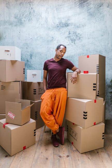 A person dressed in a light blue shirt is seated at a desk involved in a home relocation project, surrounded by various packing and moving materials. They are using a laptop displaying colorful lines 
