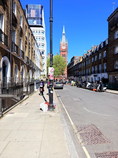 A street scene in Brixton showing terraced residential buildings with brick facades and white window frames on both sides of the road, under a clear blue sky. The sidewalk features a black metal fence